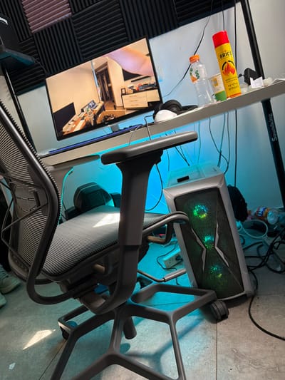 This image captures an unoccupied computer workstation situated within a room in Migdal HaEmeq, Israel.
The scene is dominated by a grey ergonomic office chair with a mesh seat and backrest, positioned in the foreground, facing a white-topped desk supported by dark metal legs, one of which bears "Soundking" branding. No individuals are present in the frame.
On the desk, an LG brand monitor displays an indoor room scene, appearing to be a reflection or a real-time camera feed of another workspace with a desk and computer setup. A keyboard is positioned in front of the monitor. To the right of the monitor, a pair of white over-ear headphones rests on the desk. Further right, a clear plastic bottle with an orange cap, containing a small amount of orange liquid, stands next to a clear plastic cup with Hebrew text. A red and yellow aerosol can, labeled "BRICO" and "WOOD LIGHTER / חומר בעירה לעץ" (Wood Kindling / Firelighter for Wood) in Hebrew, is also present. Various cables are visible on and behind the desk.
Under the desk, blue LED lighting illuminates the area. A desktop computer tower, white with grey accents and featuring three illuminated green circular fans on its front panel, rests on the tiled floor to the right of the chair. The floor around the computer tower and under the desk is cluttered with a tangle of black cables, power strips, and several discarded plastic bottles, one identifiable as Fanta. The wall behind the monitor is covered with dark grey acoustic foam panels. Natural light casts a beam onto the seat of the office chair.