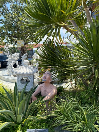In the city of Tuy An Nam, Vietnam, a garden scene unfolds under a bright sky. A statue of an elderly man with graying hair and beard sits amongst lush greenery. The man is depicted with his eyes closed and a serene expression, his chest bare. He appears to be leaning on something. In the background, a stone pagoda-like structure with dragon carvings stands tall. A car is visible parked to the left, and a yellow sign with "WC" is attached to a tree. Palm trees and other foliage create a dense, verdant atmosphere.