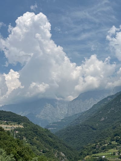 This media file captures a view of a mountainous landscape under a bright, partially cloudy sky. The scene is dominated by a steep, heavily forested mountainside that slopes down into a valley. The trees are a deep green, suggesting a time of year with lush vegetation, likely spring or summer.

Scattered across the mountainside are small buildings and structures, indicative of a rural or semi-rural settlement. Some appear to be houses, while others might be agricultural buildings or part of a small village. Power lines or cables are visible stretching across the mountain, suggesting infrastructure connecting the area.

In the distance, rugged, rocky mountain peaks can be seen, some shrouded in a light haze, hinting at atmospheric perspective and the vastness of the terrain. The sky is a mix of bright blue and large, fluffy white clouds, with some thinner, wispy clouds also present. The lighting suggests it is daytime, likely mid-morning to afternoon, with the sun illuminating the scene.

The overall impression is one of natural beauty combined with human habitation integrated into a dramatic mountainous environment. The location context of Malegno, Italy, points to a region in the Alps, known for such landscapes. No people are visible in the frame, and there is no discernible text.