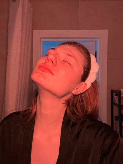 The image depicts a female individual indoors in Nikol'skii, Russia. The subject, with light-colored hair, is shown from the chest up, with her head tilted sharply backward, eyes closed, and chin elevated. Her lips are slightly parted. She is wearing a dark, smooth-textured robe. A light-colored, textured headband secures her hair. The scene is illuminated by a strong reddish-orange light source, casting a deep red hue across the subject's face, neck, and the immediate surroundings. To the left of the subject, a vertical light-colored curtain is present. Behind the subject's right shoulder, a white doorframe is visible, through which a light-blue illumination emanates. A dark, multi-tiered shelving unit is partially discernible on the far right.