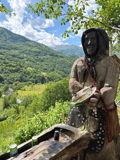 The image depicts a mountainous landscape in Dragovića Polje, Montenegro, featuring a carved wooden statue functioning as a water fountain in the foreground.
In the immediate foreground, a weathered wooden statue of an elderly figure, likely a woman, with a hooded head is positioned. The statue's torso is adorned with numerous small coins hammered into the wood, and a red and black braided rope or strap drapes over its left shoulder, extending to a small, woven fabric pouch on its right side. A metal spigot emerges from the statue's chest area, directing water into a shallow, carved wooden basin held by the statue's hands. This basin features a green, leaf-shaped extension that channels the water into a larger, rectangular wooden trough situated below. Within this trough, which contains flowing water, a Fanta Tropical soda can, another partially visible green can, and a dark-colored bottle with a white cap are placed, presumably for cooling. A small, light blue/green object rests on the statue's upper right chest.
The midground reveals steep, verdant mountainsides densely covered with deciduous trees and lush vegetation. Scattered across these slopes are several small houses with brown roofs. In a valley clearing, power lines with supporting poles are visible.
The background features more distant, higher mountain ranges with varying shades of green foliage and some visible rocky areas. The sky above is bright blue with scattered white cumulus clouds. Tree branches with green leaves partially frame the top of the image. The scene is illuminated by natural daylight. No human figures are present.