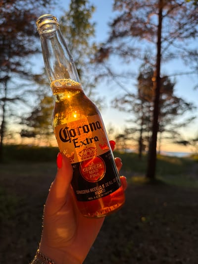 In Meremõisa, Estonia, a hand with bright red nail polish holds a glass bottle of Corona Extra beer at sunset. The bottle is tilted, with the golden liquid catching the last rays of sunlight, creating a warm glow. Bubbles are visible near the neck of the bottle, and the label, featuring the Corona Extra logo and a crown, is clearly legible. The background is blurred, showing tall trees and a hint of the sea in the distance, bathed in the soft light of dusk. The overall impression is one of a relaxed evening outdoors.