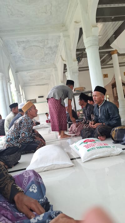 In a covered outdoor area, possibly a mosque or community hall, in Grong-grong, Indonesia, a group of men are gathered, seated on a white tiled floor. The men are dressed in traditional Islamic attire, including peci caps and sarongs. One man, wearing a grey shirt and a patterned maroon sarong, stands and appears to be interacting with the seated individuals. In the foreground, two large white sacks, presumably containing rice, are visible, with one sack displaying Indonesian text that reads "BERAS MUTU TERJAMIN". The architectural style features white columns and ornate white ceiling decorations, suggesting a well-maintained structure. The lighting appears to be natural, indicating daytime. The overall scene depicts a communal gathering, possibly for a religious event or distribution of aid.