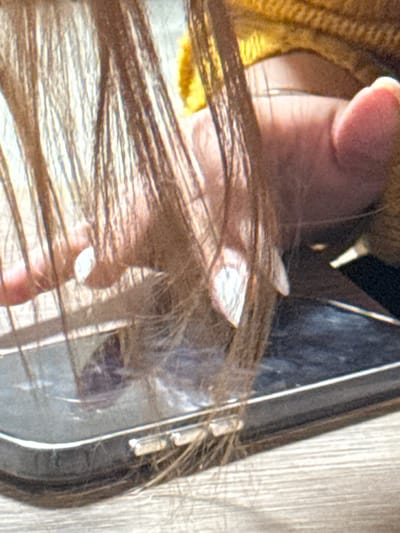 A close-up shows a hand holding a pair of glasses. The glasses have a dark frame and a clear lens. The lens reflects light. Strands of brown hair overlap the hand and the lens, creating a layered effect. A hint of yellow material is visible behind the hand. The background has a light-colored wooden surface.
