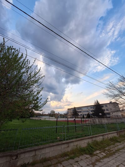 This image captures an outdoor scene in Dolni Chiflik, Bulgaria, depicting a mix of natural elements and urban structures, likely during late afternoon or early morning due to the lighting.
The sky dominates the upper portion of the frame, displaying a dynamic mix of clear blue patches, fluffy white clouds, and darker grey clouds. Several dark utility cables or power lines stretch diagonally across the sky, adding a linear element to the composition.
In the mid-ground, a multi-story building, possibly an apartment complex or a school, is visible with numerous windows. In front of the building lies a green field, which appears to be a sports ground given the visible red track surface and what looks like a basketball hoop structure. A metal fence separates this green area from a paved walkway or path made of concrete slabs. Grass and weeds grow along the edges of the path and between the slabs.
The foreground on the left features a vibrant green bush or small tree with new foliage, indicating spring or early summer. Another tree with bare branches is visible towards the top right, near the building.
There are no people or discernible activities taking place, presenting a tranquil, static view of the environment. No text is visible within the image.