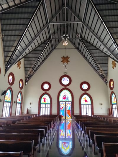 This is an interior shot of a church, likely located in K'Rèn, Vietnam, with rows of dark wooden pews facing a central altar area. The ceiling is high and vaulted, constructed with corrugated metal roofing supported by exposed metal beams. The walls are white, and the floor is highly reflective, showing the colorful stained-glass windows and the stained-glass doors at the front.

The front of the church features large, arched stained-glass windows and double doors depicting religious figures. Smaller, circular stained-glass windows are positioned higher up on the side walls. Decorative wooden stars hang from the ceiling near the front. The lighting appears to be a combination of natural light filtering through the windows and overhead light fixtures.

There are no people clearly visible in the pews. A single person is visible in the distance near the front of the church, seated behind the pews on the right side. The overall atmosphere is one of solemnity and quietude.