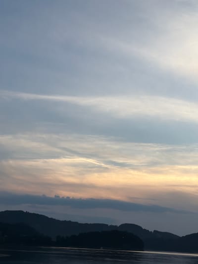 The image depicts a serene landscape at dusk. The sky is filled with soft, wispy clouds, transitioning from a pale blue at the top to a warm, golden hue near the horizon. Below the sky, dark, silhouetted hills form a gentle curvature across the lower portion of the image. The foreground is occupied by a body of water, reflecting the muted colors of the sky and the dark forms of the hills. The overall impression is one of tranquility and natural beauty. The location is Gródek nad Dunajcem, Poland.