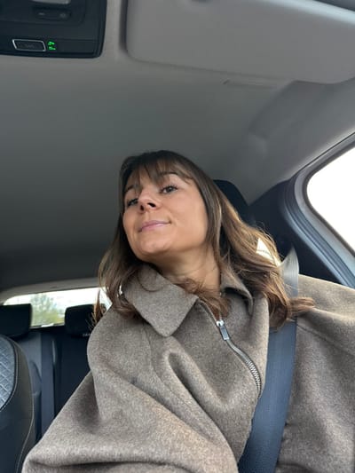 Inside a vehicle, a woman with brown hair and bangs wears a gray coat and a seatbelt. She looks at the camera with a subtle smile. The car's interior is visible, including the roof, window, and control panel. The seat is black leather. Outside the window, green foliage can be seen.