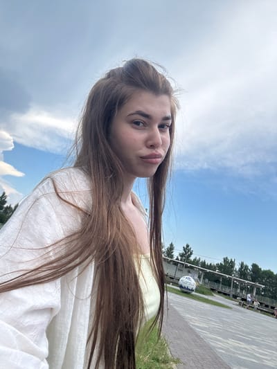 A young woman with long, light brown hair is captured in a selfie-style shot, looking directly at the camera with a slightly pursed expression. She is wearing a light yellow top and an open white linen shirt draped over her shoulders. The background suggests an outdoor setting on a partly cloudy day with a bright blue sky. In the mid-ground, a paved walkway is visible, lined with brick. Further in the background, there is a structure resembling a building or pavilion with a covered walkway, surrounded by trees. A large, decorative spherical object, possibly a globe, is placed on the ground near the structure. Several people are seen in the distance, some walking and others sitting on benches, indicating a public space. The overall impression is of a casual outdoor excursion in a park or waterfront area. While the image does not explicitly show any landmarks that pinpoint it to the city of Речицы, Russia, the presence of a public recreational area with trees and paved walkways is consistent with park environments found in many cities.