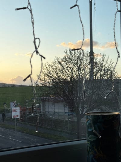 The image depicts an outdoor scene in Vellmar, Germany, viewed through a window, possibly at dusk or dawn. In the immediate foreground, out-of-focus string lights with individual bulbs hang across the upper and central portions of the frame, and a decorative cylindrical object, possibly a mug or candle holder with an ornate pattern, is partially visible in the bottom right corner.
Beyond the window, a paved road traverses the lower-left, bordered by a sidewalk to its left. A single person, dressed in dark attire, walks leftwards on this sidewalk. Adjacent to the road, a yellow diamond-shaped traffic sign and a red and white political advertisement sign for "Die Linke" are visible, bearing the German text "Kümmern statt Kären."
A construction site occupies the midground, featuring scaffolding surrounding an incomplete structure or building, enclosed by a wire fence. A large, deciduous tree with bare branches dominates the right-center, positioned in front of the construction area. Behind the tree, a tall, slender pole extends vertically into the sky.
The background on the left consists of green fields and distant rolling hills. The sky presents a gradient of light blue, transitioning to pale yellow and orange near the horizon, indicating low sun conditions. Scattered white and light orange clouds are present in the upper sky.