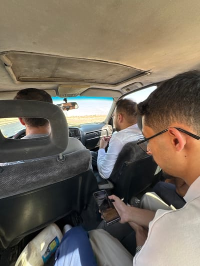 The image displays the interior of a moving vehicle, captured from a rear-seat perspective looking forward. Four adult males are present within the vehicle. The driver is positioned in the front left seat with hands on the steering wheel. A male front-right passenger, wearing a white long-sleeved shirt and glasses, is seated with hands resting on his lap. In the rear right seat, a male passenger with dark hair and black-framed glasses holds a black smartphone horizontally with both hands, viewing its screen. He wears a light-colored shirt and a watch on his left wrist. His legs, clad in light-colored trousers, are visible alongside a leg in dark blue trousers. In the rear left footwell, a leg in dark blue trousers and white footwear is visible next to a large, light-colored cylindrical object. The vehicle's interior features a light grey, worn headliner with a closed sunroof and dark grey fabric seats. The exterior environment, visible through the windshield, consists of a paved road traversing a light brown, arid landscape under a clear blue sky. This scene is located in the City of Adra labor, Syria.
