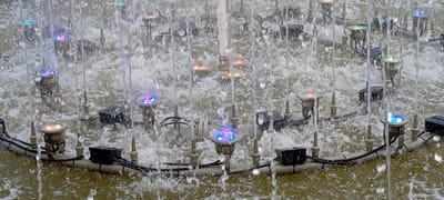 The image depicts a close-up view of a water fountain with multiple illuminated nozzles spraying water upwards. The fountain is situated outdoors, with the water appearing somewhat murky, suggesting it may be in a pond or basin. The nozzles are arranged in a semi-circular pattern, and each is equipped with a colored LED light, displaying a variety of hues including blue, pink, green, and red. The water jets create a dynamic, abstract pattern of streaks and droplets against a textured background of agitated water and submerged structures. Some nozzles are more brightly lit than others, and the lights refract and reflect off the water, adding to the visual complexity of the scene. The overall impression is one of a decorative and lively water feature.