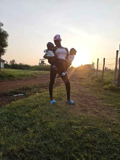 The image depicts an adult female holding two infants outdoors at sunset in Ang'orom ward, Kenya. The adult, positioned centrally on a reddish-brown dirt path with green grassy verges, wears a white tank top, dark leggings, a red and white patterned head covering, and light blue slide sandals. She holds one infant, dressed in a white top and light blue shoes, in her left arm. A second infant, wearing a light-colored long-sleeved shirt, is held in her right arm. In the left background, a small white building with a dark roof is partially visible behind a tree, with a water tank on a tower structure further in the distance. To the right, a wire fence with wooden posts extends towards the setting sun on the horizon, which casts a warm, golden light across the scene and creates some lens flare. The immediate foreground consists of dense green grass.