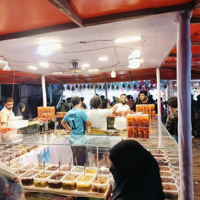 This media file captures a bustling food stall scene, likely a marketplace or fair, at night, given the reliance on artificial lighting. The location is Charfasson, Bangladesh.

The main subject is a brightly lit stall featuring a variety of food items, predominantly different kinds of pickles or chutneys (achar). These are displayed in numerous clear plastic containers arranged in rows on and behind a glass counter. Many small, transparent jars filled with reddish, possibly dried fruit or chili-based snacks, some labeled "Natural," are stacked on the counter.

Several people are visible in the image. In the foreground, a person in a black hijab is partially obscured, looking intently at the food displays. Behind the counter, there are multiple vendors and staff interacting with customers. On the left, a man with a beard and a woman in a black hijab are present. In the center, a man wearing a blue t-shirt with "AMIRUL 11" printed on the back faces away from the camera, conversing with another man with curly hair. On the right, two men are smiling, one in a white shirt and another in a black shirt, engaging with customers. Blurred figures in the background indicate a lively crowd.

Overhead, a red canopy shades the stall, from which multiple fluorescent tube lights and incandescent bulbs hang, along with a small fan. Exposed electrical wires are visible. The background shows corrugated metal sheets and colorful, possibly decorative, plates or objects hanging. The overall atmosphere appears vibrant and active. A sign at the bottom of the counter, partially visible, displays Bengali script: "বিসমিল্লাহির রাহমানির রাহিম" (Bismillahir Rahmanir Rahim), meaning "In the name of Allah, the Most Gracious, the Most Merciful."