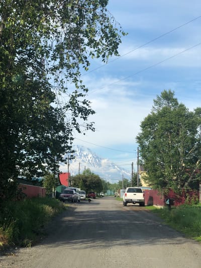 A dirt road leads into the distance, lined with houses and trees. A large snow-capped mountain is visible in the background, under a partly cloudy sky. Several vehicles are parked along the road. A dog walks in the middle of the road.