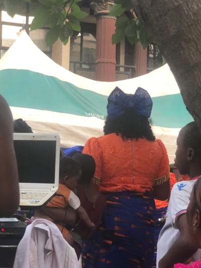 The scene, located in Awka Etiti, Nigeria, depicts an outdoor gathering, possibly an event or market. In the foreground, a large tree trunk with green foliage frames the right and top portions of the image. The central figure is a woman viewed from behind, wearing a textured orange short-sleeved top with gold embellishments on the sleeves, and a blue wrapper skirt featuring a geometric pattern. Her dark hair is styled with a prominent blue bow. To her immediate left, an adult's arm cradles a partially visible infant or small child, who wears an orange and white garment. A white laptop with an open black screen is visible on a surface to the left of the child, near which white fabric is draped. To the right of the central woman, a young boy in a white short-sleeved shirt with a blue logo on the chest is partially visible. In the mid-ground, a large white and light blue striped tent, partially displaying the word "LUCKY," serves as a backdrop. Behind the tent, parts of a building with brown columns and windows are visible, indicating an urban setting. The overall impression is a vibrant public assembly.
