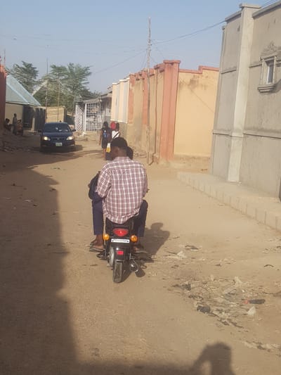 A street scene in Dutsinma A, Nigeria, features a person on a motorcycle, oriented perpendicularly to the road. The individual, wearing a plaid shirt and dark pants, is seated on the motorcycle. Nearby, a blue car is parked. Other people are visible in the distance. The road is unpaved, and there's debris scattered on the ground. Buildings line one side of the road. The sky is clear and light blue.