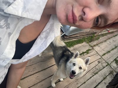 An upward-angled, selfie-style photograph captures a person and a dog standing on a wooden deck outdoors. The person, positioned in the upper left of the frame, is wearing a white floral-patterned shirt. Only the person's chin, mouth, and a portion of their face are visible, turned towards the camera. The dog, a husky mix with heterochromatic eyes (one brown, one blue), is in the center of the frame, looking up at the camera with its mouth open. The wooden deck appears weathered, with visible gaps between the planks and patches of grass growing through. In the background, a metal bucket and some stacked wooden planks are discernible. The scene suggests a casual outdoor setting, possibly a backyard or a rural location, consistent with areas outside of Mezhdurechenskiy, Russia, where such natural and unmanicured environments are common.
