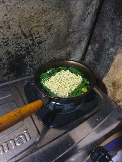 The scene depicts an instant noodle dish being cooked on a gas stove indoors in thành phố Biên Hòa, Vietnam. A dark-colored pan with a worn, light brown handle rests on a silver Rinnai gas stove, with a visible blue flame heating its contents. Inside the pan, a block of dry, yellow instant noodles floats atop green leafy vegetables, submerged in a dark liquid. The background consists of a dark, textured wall exhibiting discoloration and wear. No individuals are visible.