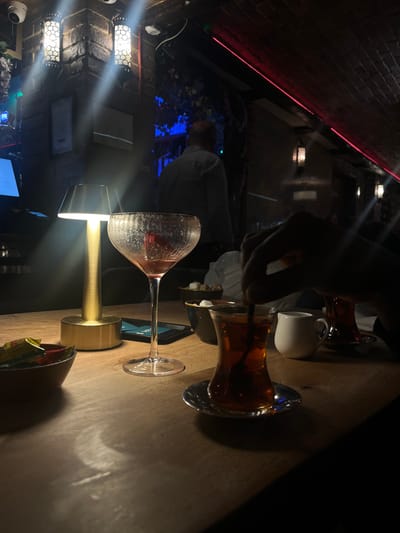 The image displays the interior of a dimly lit establishment, possibly a lounge or restaurant, during evening hours. The specific location within Stevenage, United Kingdom, is not discernible from the visual information provided.
In the foreground, a wooden table surface holds multiple items. From left to right: a small, dark wooden bowl containing individually wrapped candies or sugar packets with red and yellow wrappers; a gold-toned, illuminated portable table lamp emitting a bright light beam; a clear, ridged cocktail glass containing residual reddish liquid; a small, dark bowl with white sugar cubes; a dark-colored smartphone, partially illuminated; a pear-shaped Turkish tea glass on a metallic saucer, filled with dark amber liquid, being stirred by a dark-skinned hand holding a spoon; and a small white ceramic creamer.
The background features a dark, textured wall, partially constructed of exposed brickwork, adorned with two ornate, geometric-patterned wall sconces casting light upwards. A section of the wall to the left appears covered in dark artificial foliage. A red LED strip light runs along the upper right ceiling line. A blurred male figure, with short or no hair, wearing a light-colored shirt, is visible from the back, positioned away from the camera. A white dome security camera is mounted high on the left wall. Distant blue ambient lighting is visible in the upper left background.