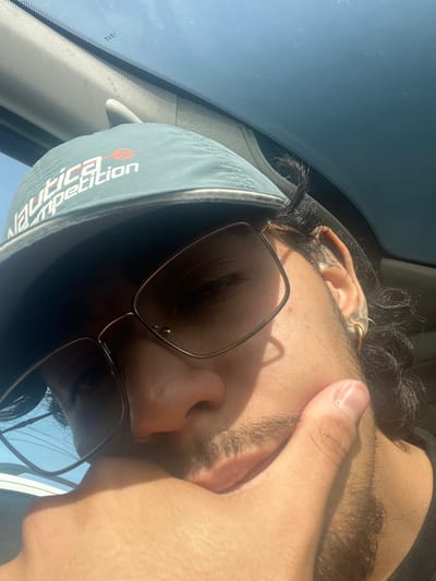 This image captures a close-up, low-angle selfie of an adult individual inside a vehicle in Tolhuin, Argentina, under bright daylight conditions. The subject wears a grey Nautica Competition baseball cap with a red and white logo, along with rectangular, dark-lensed sunglasses. Dark, wavy hair is visible around the right ear, which features a metallic hoop earring in the lobe and a tattoo on the cartilage. Facial stubble is present on the chin and jawline. The individual's right hand, with visible thumb and index finger, rests against their chin or lower jaw. Strong sunlight illuminates the scene, casting distinct shadows on the face and hand, and creating reflections on the sunglasses and the vehicle's windshield. Visible vehicle interior components include a grey A-pillar trim and the upper section of the windshield, through which a clear blue sky is observed. No other individuals or significant interactions are discernible.