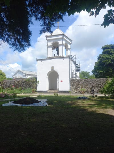 The image depicts the bell tower of a white building, likely a church, located in Ilori, Abkhazia. The bell tower is square with arched openings, topped with a pointed roof and a cross. A spiral staircase is attached to the side of the tower. In the foreground, a pile of dark material, possibly soil or compost, sits on a white tarp spread on a grassy area. A stone wall runs along the perimeter of the property, separating it from a road where a bicycle is parked. Trees and foliage surround the building, with a clear blue sky and scattered clouds above. The scene appears to be outdoors on a sunny day, possibly in a rural or semi-rural setting within Ilori.