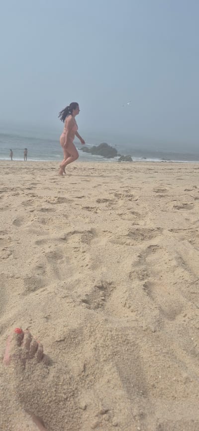 In Póvoa de Varzim, Portugal, a woman in a pink bikini walks along a sandy beach. Her hair is pulled back in a ponytail, and she is in motion, with her left leg extended forward. The beach is covered in sand, with visible footprints. In the background, the ocean meets the shore, with waves crashing against rocks. The sky is hazy and overcast, with a seagull flying in the distance. Two children are visible in the shallow water near the shore. In the foreground, a person's foot, with red-painted toenails, is partially submerged in the sand.