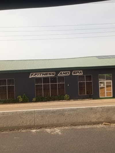 This is a medium-shot, eye-level photograph of a building with the text "F. FITNESS AND SPA" on its exterior. The building has a dark grey facade and a green, corrugated metal roof. There are several large windows with dark tinting and metal bars. To the right, there is a glass door with a white frame. In front of the building, there is a line of manicured bushes and small trees. The building appears to be situated in a relatively open area, with a road and a concrete barrier in the foreground. The sky is overcast and light, suggesting a cloudy day. The overall impression is of a commercial establishment, likely a gym or fitness center. The image quality is decent, with the text clearly legible.