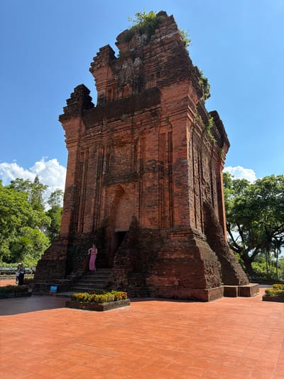 This is a full-shot, eye-level photograph of the Thap Nhan, a Cham tower located in the city of Tuy Hoa, Phu Yen province, Vietnam. The ancient red brick tower, adorned with greenery on its upper sections, dominates the center of the frame. The tower exhibits a stepped pyramid shape with a prominent entrance. To the left of the tower, two women are visible: one in a pink dress stands on the steps leading to the entrance, and another, wearing a dark top and light-colored pants, stands further back on the paved ground. The foreground is covered with terracotta tiles, and in front of the tower's base are rectangular planters filled with small red flowers. The background features lush green trees and a bright blue sky with scattered white clouds.