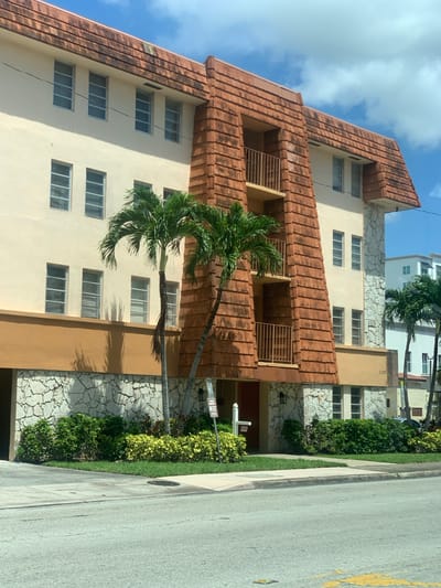 The image shows a building located at 1700 on a street in Coral Gables, Florida, United States of America. The building is a multi-story structure with a tan facade and a distinctive tiled roof that appears to be made of clay tiles. A central section of the building features a textured, reddish-brown facade, possibly concrete or stucco, with balconies. The lower part of the building is clad in irregular, light-colored stone. Two palm trees are situated in front of the building. The ground level has greenery and a sidewalk. A street with a yellow painted line is visible in the foreground. The sky is blue with scattered clouds. The overall impression is of a commercial or residential building in a sunny, possibly tropical, environment.