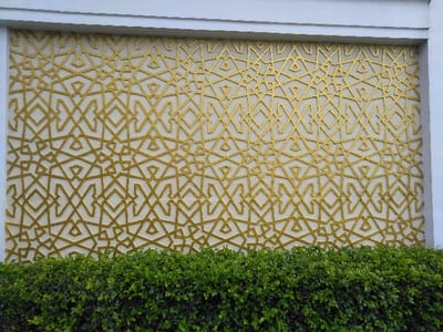 The image displays a decorative architectural panel featuring an intricate golden-yellow geometric pattern set against a light, possibly off-white or cream, background. The pattern is composed of interconnected lines forming repeating abstract shapes, including rhombuses, squares, and angular configurations, arranged in a tessellated design. This patterned section is framed by a plain white border at the top and sides. A dense, verdant green hedge occupies the lower portion of the frame, obscuring the base of the wall and extending horizontally across the entire width of the image. There are no individuals, animals, or other discernible objects present in the scene. The location is specified as the city of San Cristóbal, Costa Rica.