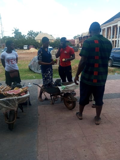 This image depicts an outdoor scene in Ajalli, Nigeria, likely a street or market area. Four individuals are present on a reddish-brown patterned paved surface, with two wheelbarrows in the foreground.
The leftmost wheelbarrow, positioned on a dirt patch, is a vendor's stall, laden with cut fruits including watermelon slices and possibly pineapples, alongside what appears to be groundnuts, partially covered by a translucent sheet. Adjacent to it, a second, mostly empty rusty wheelbarrow with a white plastic bag is situated on the paved area.
Four individuals are engaged in an interaction:
1. A woman on the far left, wearing a white t-shirt with "YES" in red text and a dark skirt, stands with her hands clasped in front of her.
2. Next to her, another woman in a dark blue floral dress, barefoot, holds a green, round fruit in her hands. She faces towards the right.
3. A man in a red polo shirt, dark trousers, and a black baseball cap is actively consuming a light-colored fruit, holding it to his mouth, with another similar fruit in his left hand. He faces the two women.
4. On the far right, a man with his back mostly to the camera wears a dark green and black striped polo shirt with red vertical rectangles on the back, dark shorts, and brown flip-flops. He faces the other three individuals.
The background shows a mix of urban and semi-urban elements under a bright sky. To the left, a tall metal antenna tower rises above green foliage and several vehicles in a dirt area with piles of construction materials. To the right, a large multi-story building with white pillars and a dark roof is visible, along with another building under construction and a parked dark blue SUV.