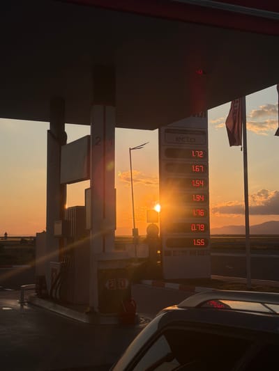 The image shows a gas station with a sign displaying fuel prices: 1.72, 1.67, 1.54, 1.94, 1.76, 0.79, and 1.55. The gas station is branded with "ecto" and the words "ИНОВАЦИОННИ ГОРИВА." A gas pump is visible, labeled with the numbers "2" and "1." A car is partially visible on the right side of the frame. The background shows a landscape with a setting or rising sun, casting a strong light and creating silhouettes. The location is Vodenichane, Bulgaria.