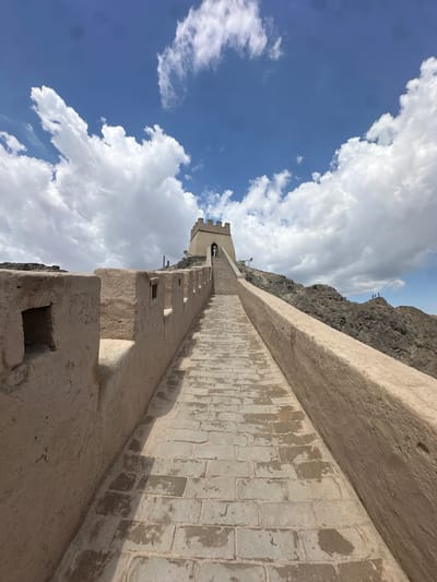 The image captures a section of the Great Wall of China near Jiayuguan, China. The wall, constructed from tan-colored bricks and stone, ascends a rocky, mountainous terrain under a bright blue sky dotted with fluffy white clouds. The perspective is looking up a stone-paved walkway that follows the contours of the wall, with crenellated battlements lining the left side.
A watchtower stands at the crest of the incline, its stone structure visible against the sky. Several individuals are present on the walkway and near the watchtower, appearing to be tourists. In the distance, on a rocky outcrop to the right of the wall, a small group of people can be seen. The environment is dry and arid, characterized by the rocky, rugged landscape. The lighting suggests it is daytime, with distinct shadows cast by the wall and its features on the walkway.