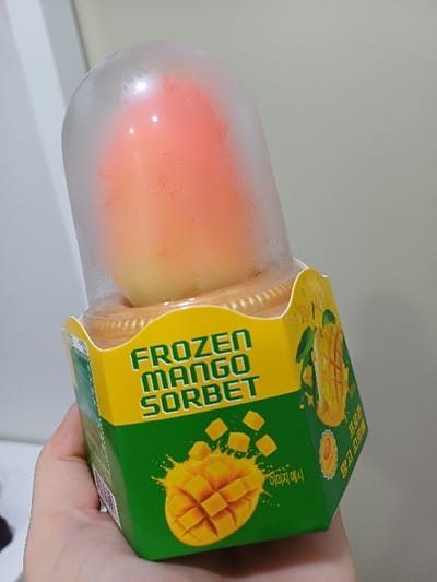 A single hand holds a "Frozen Mango Sorbet" product in an indoor setting. The product is housed in a hexagonal paperboard container with a green base and a yellow top band. Green text on the yellow band prominently displays "FROZEN MANGO SORBET." The green section features an illustration of a diced mango splashing and Korean text. A clear, dome-shaped plastic lid covers the top, revealing the two-toned, multi-layered sorbet inside, which transitions from a lighter orange-yellow at the base to a reddish-orange at the top. Condensation is visible on both the interior and exterior of the plastic dome, indicating the product's cold temperature. The hand, visible from the wrist down, has a light skin tone and holds the product from the lower left side. The background consists of a plain, light grey or off-white wall, with a subtle vertical line on the far left suggesting an architectural feature like a door frame or wall corner. The specific location within Seodaemun-gu, South Korea, cannot be determined from the visual information provided in the image.