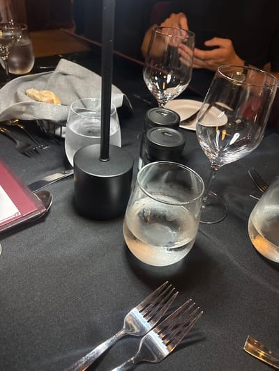 The image depicts a close-up, low-light view of a dining table set at a restaurant in Spanish Town, British Virgin Islands. The table is covered with a black tablecloth. In the foreground, two silver forks rest parallel on the cloth. To the upper right, a clear, stemless glass tumbler containing water and ice shows condensation on its exterior. Another similar tumbler is partially visible to the far right. Centered in the midground is a black, cylindrical object, likely a table lamp base or stand. To its left, another tumbler holds clear water. A gray fabric napkin cradles a piece of bread inside a dark wire basket. Two empty stemmed wine glasses stand on the right side of the table, with two black, cylindrical shakers positioned behind them. A maroon-colored menu or check holder is partially visible on the far left. In the blurred background, the hands of at least one person are visible, holding a dark, rectangular object, indicating a diner seated at the table. The environment is consistent with an indoor restaurant setting.