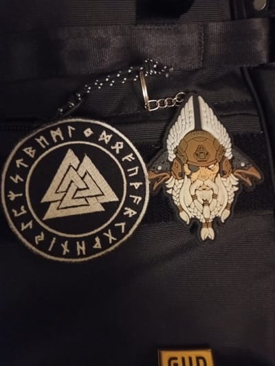 Two patches are affixed to a black fabric surface. The patch on the left is circular and embroidered with a white triple-triangle symbol (Valknut) within a circle of white runes on a black background. The patch on the right is a PVC patch depicting a stylized Viking-like character with a helmet, beard, and wings, with two black birds perched on either side of its head. A keychain with a silver chain and a black and white cord is attached to the fabric near the top center. The black fabric surface appears to be a backpack or bag. A small gold-colored metal plate with the letters "GUD" is visible at the bottom center of the image. The scene is indoors with artificial lighting. The location of the image in Sloviansk, Ukraine, cannot be determined from the visual information provided.