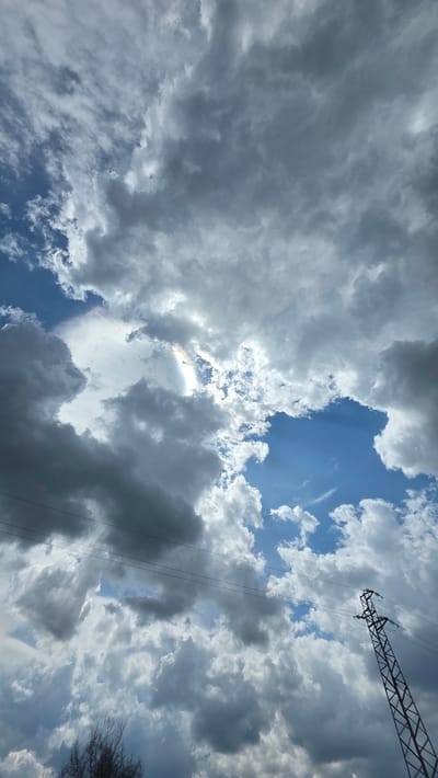 The image captures a dramatic sky filled with a mix of dark and bright clouds, looking upwards towards the zenith. The dominant subjects are the expansive sky and various cloud formations. Large, dark grey cumulus or stratocumulus clouds cover much of the frame, interspersed with patches of bright blue sky. The sun is partly visible behind the clouds, casting intense rays of light and creating a bright, almost ethereal glow.
A notable natural phenomenon is visible: a circumhorizontal arc or iridescent cloud displaying a distinct rainbow-like spectrum of colors (red, orange, green, blue) on the lower edge of a cloud illuminated by the hidden sun, slightly to the left of the center. Beams of sunlight, also known as crepuscular rays, can be seen breaking through the cloud cover.
In the upper right portion of the image, the metal lattice structure of a high-voltage power line tower is prominently visible, extending diagonally across the sky. Associated power lines run downwards from the tower. On the far right edge of the frame, sparse, dark, bare tree branches or shrubbery are partially captured. A tiny dark speck, possibly a bird or insect, is discernible against the darker clouds in some areas.
The scene depicts daytime conditions, likely mid-day to late afternoon, given the intensity of the light breaking through the clouds. The weather appears dynamic, suggesting a transition from or to an overcast state, perhaps after a rain shower or preceding one. The setting is outdoors, likely in a rural or semi-urban environment, with the power lines hinting at human infrastructure. The location context provided is Drenkovo, Bulgaria. There is no visible text in the image.