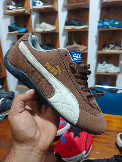 The image portrays an indoor scene, likely within a retail environment or a personal collection display in Bireuenmeunasahcapa, Indonesia. The main subject is a brown suede Puma sneaker, held by a person's hand. The shoe features a distinct white Formstrip on its side, dark brown laces, and a black sole. The brand name "PUMA" is visible in yellow text on the shoe's outer side, while a blue label or loop near the tongue displays the partial text "spo," suggesting it is part of the brand "Sparco."

In the background, numerous other sneakers of various brands and colors are arranged on light brown wooden shelves, creating a blurred depth-of-field effect. Among these, a white and black sneaker with a visible Nike swoosh is discernable on the top left shelf, and several blue and yellow athletic shoes appear on the right-side shelves. In the bottom foreground, partially visible on a wooden surface, is a red and black sneaker, also identifiable by its Puma-like formstrip. The activity centers on the act of holding and presenting the shoe, possibly for inspection or display. The setting is illuminated by artificial light, providing no specific time-of-day cues.