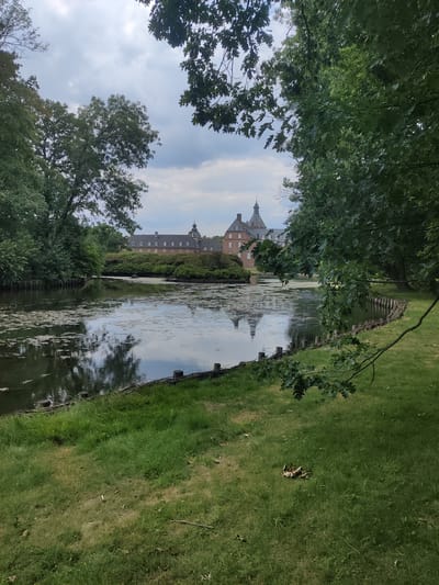 The image shows a serene landscape featuring a body of water in the foreground and a building in the background. The water, likely a moat or pond, reflects the cloudy sky and the surrounding greenery. The building appears to be a historic structure, possibly a castle or manor, with multiple windows and a distinctive tower. Lush trees frame the scene, with some branches hanging over the water, creating a sense of depth and enclosure. The ground in front of the water is covered in grass, with some fallen leaves scattered about. The overall impression is one of tranquility and natural beauty. This is a view of Wasserschloss Anholt, located in Isselburg, Germany.