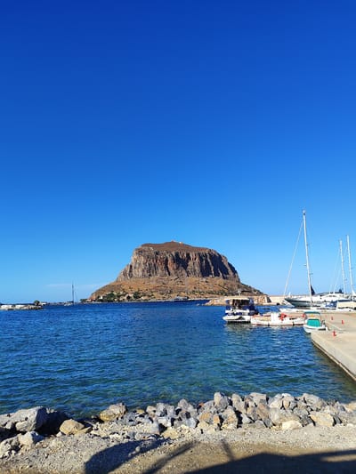 The image depicts a scenic view of the sea and a rocky island in the city of Gefyra, Greece. The foreground shows a rough stone embankment leading to clear blue water. Several boats, including sailboats and a ferry, are moored in the harbor. In the distance, a large, rocky island with a settlement and fortifications on its summit dominates the horizon. The sky is a vibrant, clear blue, suggesting a sunny day. The overall scene conveys a sense of tranquility and natural beauty.