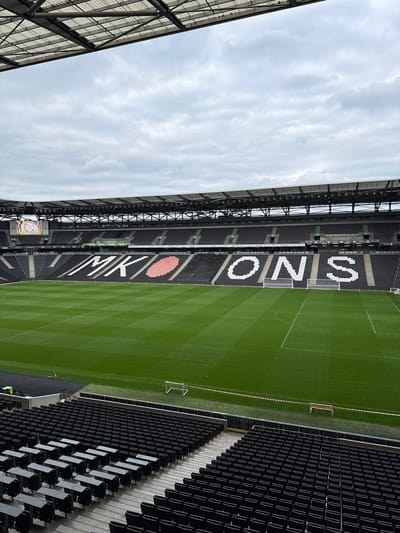 An outdoor photograph depicts a partial view of a stadium in Milton Keynes, United Kingdom, presumably Stadium MK, with a portion of the football pitch and the stands visible under an overcast sky. The grass of the football pitch is meticulously manicured with visible mowing patterns. Two goalposts stand at either end of the visible section of the pitch. The seats in the stands are predominantly black, with white block lettering spelling out "MK DONS" running vertically down the seating. A large, heart-shaped graphic is integrated into the "MK" part of the lettering. To the left, a scoreboard or large screen is visible, though the image displayed on it is indistinct. The stadium's architecture includes a complex network of metal supports and roofing. There are few people visible in the lower right corner of the image, and their actions are not clear.
