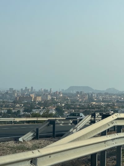The image captures a hazy panoramic view of the city of El Campello, Spain, from an elevated vantage point. In the foreground, a highway guardrail made of weathered metal and concrete is prominently displayed, with a road surface visible beneath it. A dark-colored car is seen driving on the highway. The middle ground reveals a dense urban landscape characterized by numerous buildings of varying heights and architectural styles, interspersed with patches of greenery. In the background, rolling hills and a prominent mountain range form the distant backdrop, shrouded in a light atmospheric haze. The sky above is a pale, overcast blue.