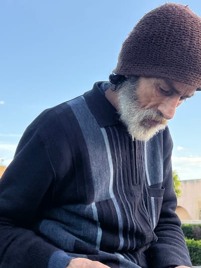 A male individual is depicted in an outdoor close-up shot within Al Machouar – Stinia, Morocco. The subject, appearing middle-aged to elderly, wears a dark brown knitted cap and a striped, collared, long-sleeved sweater. The sweater displays vertical bands of dark blue, light blue, and grey, featuring a short zippered placket and a lower right patch pocket. He possesses a substantial white beard and mustache, with darker hair visible beneath the cap. His facial expression is one of downward focus, directing his gaze below the image frame. The background is dominated by a clear blue sky. In the lower right, light-toned architectural elements and green foliage are discernible, consistent with an urban park or public garden setting.