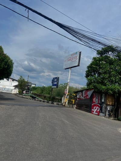 The scene is a street in Kecamatan Dayeuhkolot, Indonesia, located at latitude -6.967 and longitude 107.616. The street is paved with asphalt and slopes slightly downward towards the foreground. A sign for "PT. HARATEX" stands to the left of the road. To the right of the street is a small building with a closed metal roll-up door. The building is decorated with multiple posters and advertisements. On the left side of the street, there is a small, gated entrance. There are trees and bushes on either side of the road. Numerous utility poles line the street with power lines running overhead. The sky is a clear, light blue with white puffy clouds. There are no people or vehicles in the scene.