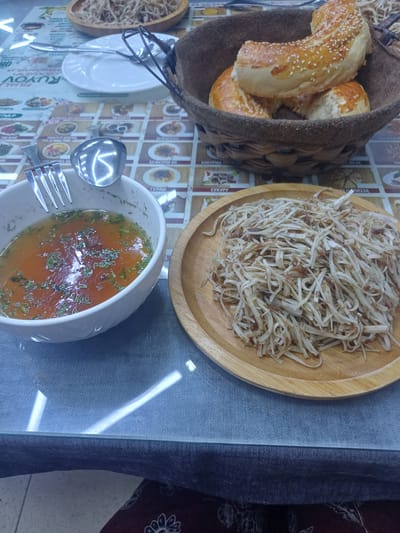 The image depicts a table setting in what appears to be a restaurant in Shahand, Uzbekistan. The table is covered with a patterned tablecloth. In the foreground, to the left, is a white bowl filled with a clear, reddish-brown broth garnished with chopped green herbs. A fork and a spoon rest on the rim of the bowl. To the right of the soup bowl is a round, light-colored wooden plate piled high with a mound of shredded noodles, possibly served as a side dish. In the background, to the right, is a woven basket containing several golden-brown, sesame-seed-covered pastries, likely samsa or similar baked goods. The overall impression is a simple meal presented on a table, possibly a cafe or a modest dining establishment.