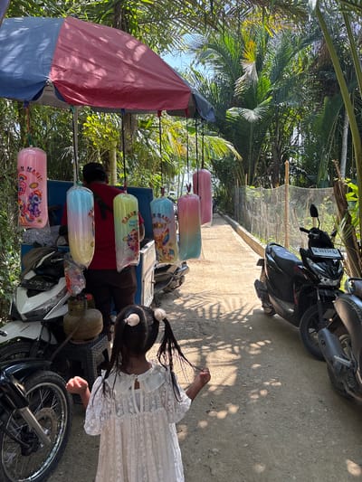 A young girl with pigtails stands on a dirt path in front of a roadside vendor's stall in Kruengseupeng, Indonesia. The stall, covered by a red and blue tarp, displays several large, colorful bags of cotton candy hanging from the awning. The cotton candy bags are decorated with cartoon characters such as My Little Pony, and other animated figures. Behind the girl and the stall, several motorcycles are parked. A man in a red shirt and black cap stands behind the stall, appearing to be the vendor. The environment is tropical, with lush green palm trees and foliage surrounding the scene. The path appears to be a common road with a fence running alongside it, indicating a residential or rural area. The sun is shining brightly, casting shadows on the ground.