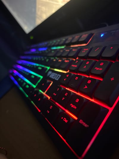 The image captures a close-up, angled view of a black computer keyboard with active multicolored RGB backlighting, located in Tsenovo, Bulgaria. The perspective is low, looking from the lower-left corner across the keyboard to the upper-right. The keyboard features a full layout, including a numeric keypad on the right. Key illumination transitions from green and blue on the far left, through purple and pink in the central QWERTY section, to predominantly red on the numeric keypad and arrow keys on the right. The brand "ARES M3" is visible on a keycap. A blue indicator light, likely for "Num Lock," is illuminated above the numeric keypad. The keys appear dark or black. The background is dark and out of focus, suggesting an indoor environment with low ambient light, with a faint, blurry light source or reflection visible in the upper background. A small, out-of-focus red and white striped object, possibly a bracelet, is partially visible in the bottom-left corner of the frame. No individuals are present, and no active human interactions or explicit actions are depicted.
