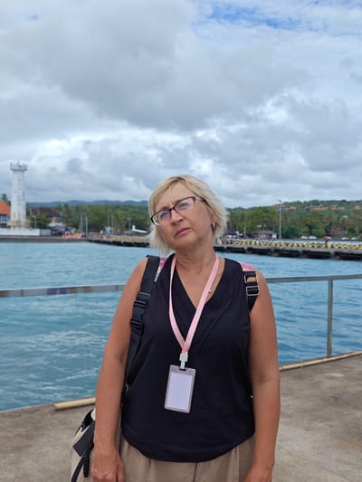 A woman stands on a pier in Kampung Toyapakeh, Indonesia, with the ocean in the background under a cloudy sky. She wears glasses, a black tank top, and a khaki skirt, with a pink lanyard around her neck. A backpack is slung over her shoulder. In the background, a white lighthouse is visible on the left, and a pier extends into the water on the right, with land and trees in the distance. A metal railing runs along the edge of the pier where the woman stands.