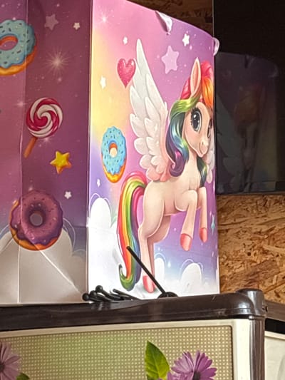 A close-up, eye-level, indoor shot shows the side of a cardboard box decorated with a unicorn and donuts. The box is pink and purple, with a unicorn with a rainbow mane and tail and white wings. There are also donuts and lollipops on the box. The box is sitting on top of a dark brown object that appears to be a piece of furniture. The background is out of focus and appears to be a wooden wall. The scene is likely indoors, possibly in a home or store. No people are visible. The objects are clean and appear to be new. The lighting is even and there are no shadows.